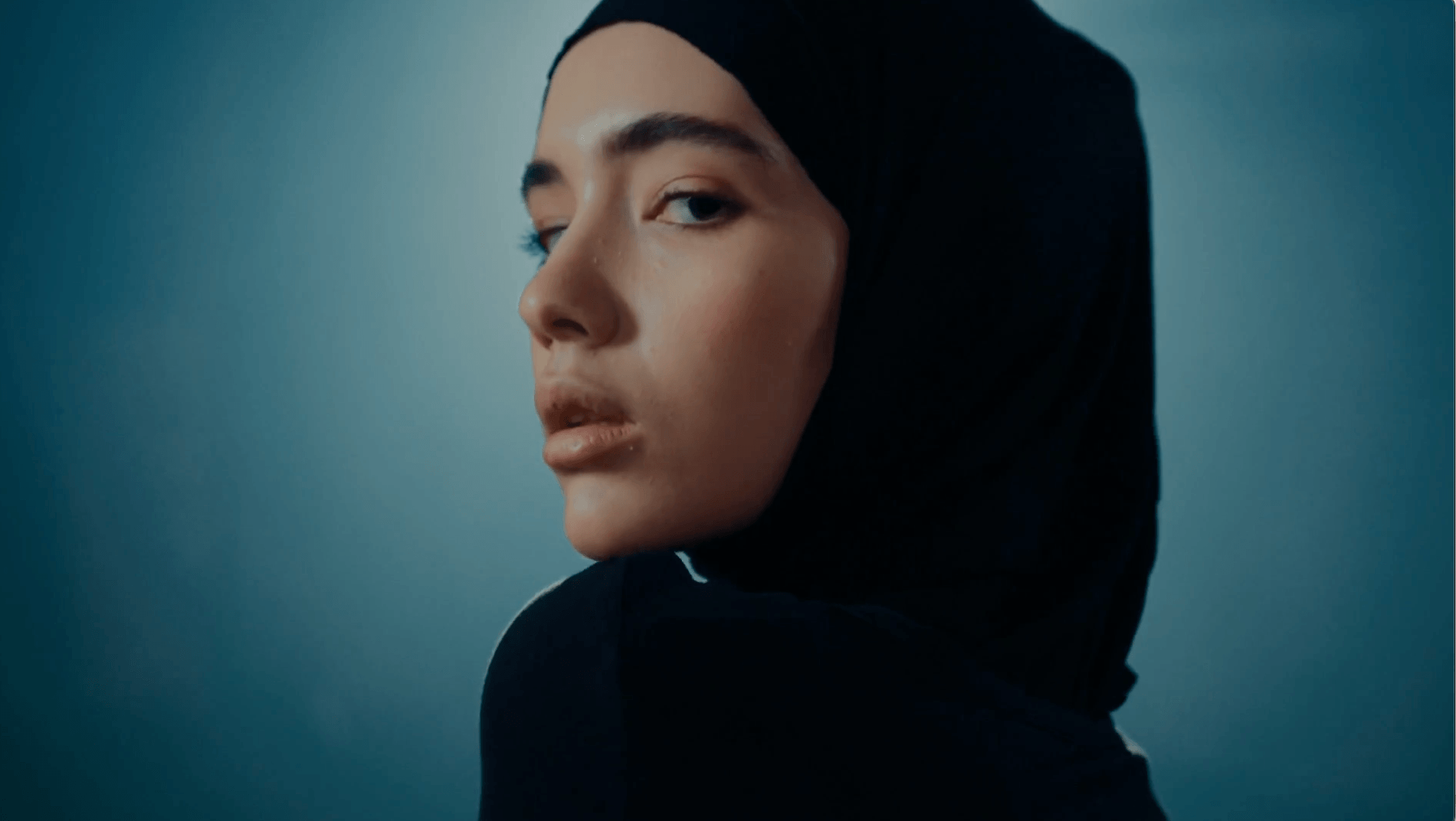 A person wearing a black hijab looks over their shoulder against a blue gradient background.