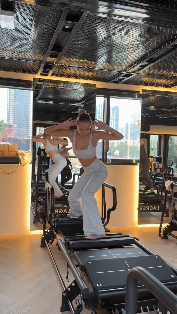 A woman in workout attire exercises on a reformer machine in a mirrored studio with city views in the background.