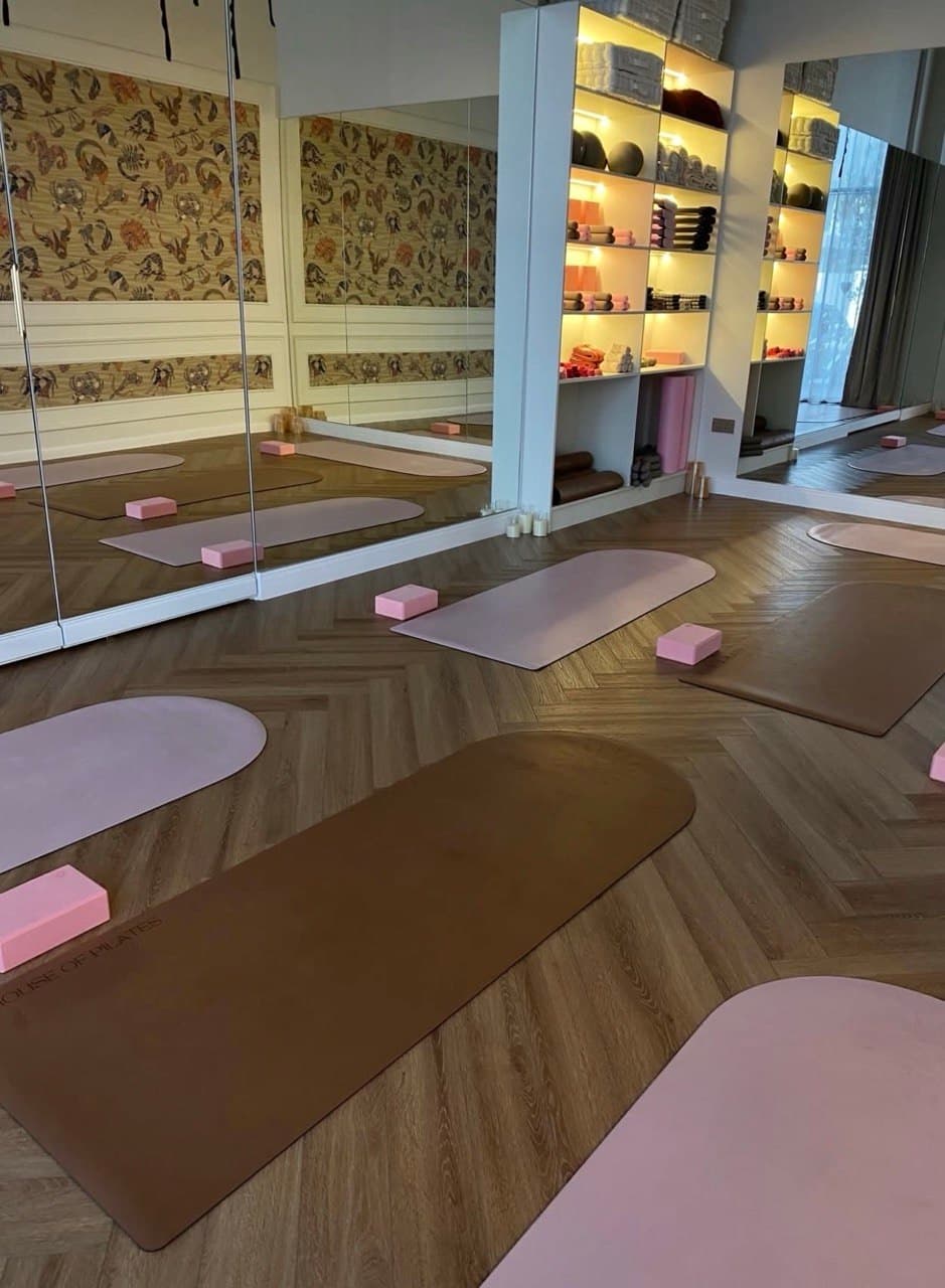 Yoga studio with pink and brown mats, pink blocks, mirrored wall, warm lighting, shelves with towels and decor, and wood-patterned floor.
