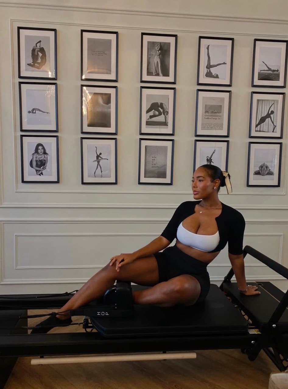 A person in workout attire sits on a Pilates reformer in front of a wall with framed black-and-white photos.