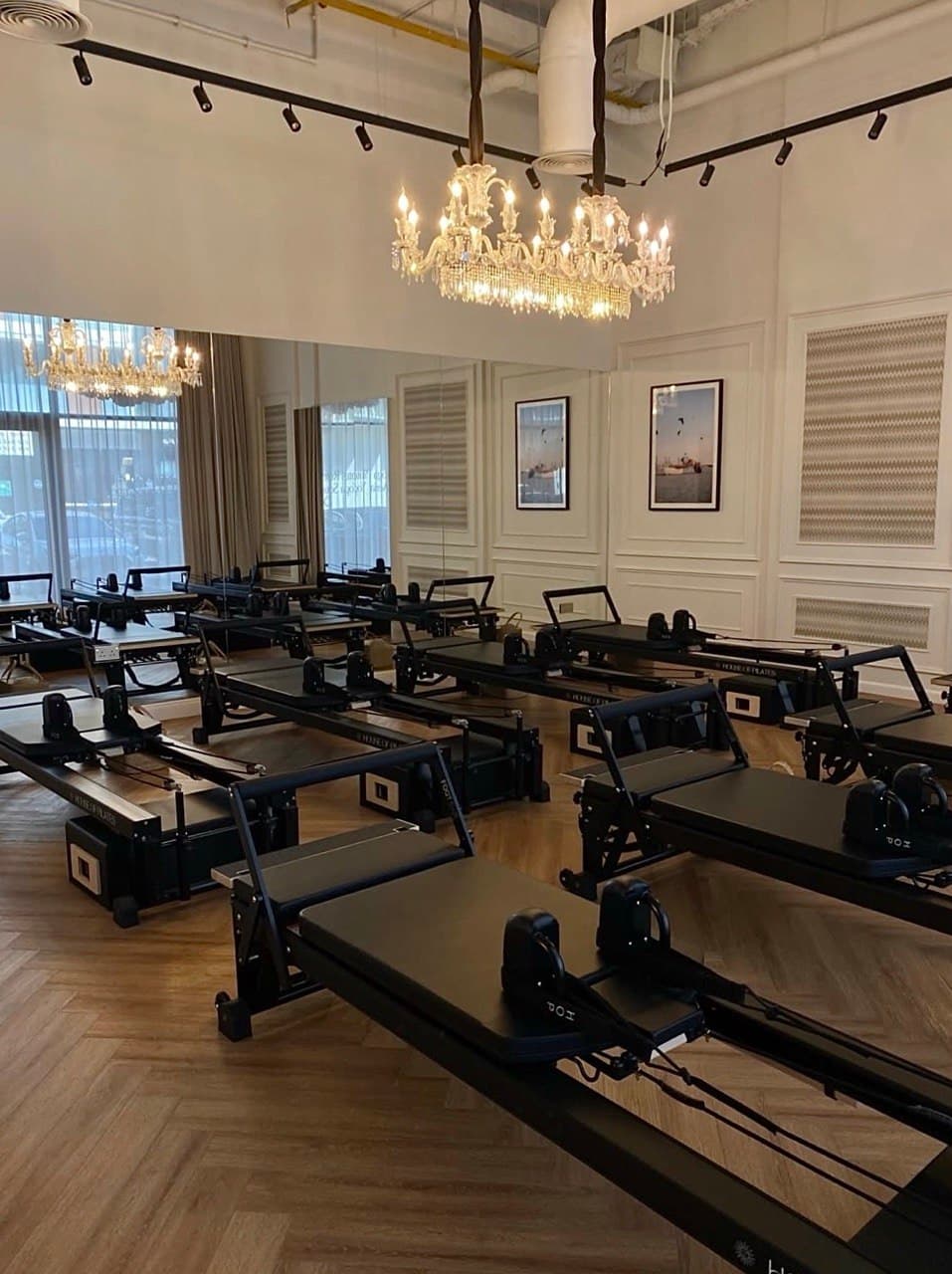A Pilates studio with multiple reformer machines, elegant chandeliers, and framed art on the walls, featuring wooden flooring.