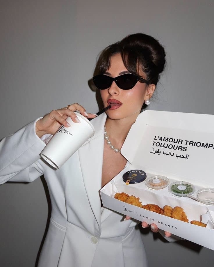 Woman in a white blazer, pearl necklace, and sunglasses holds a cup and open food box labeled "L'AMOUR TRIOMPHE TOUJOURS," with dips and snacks.