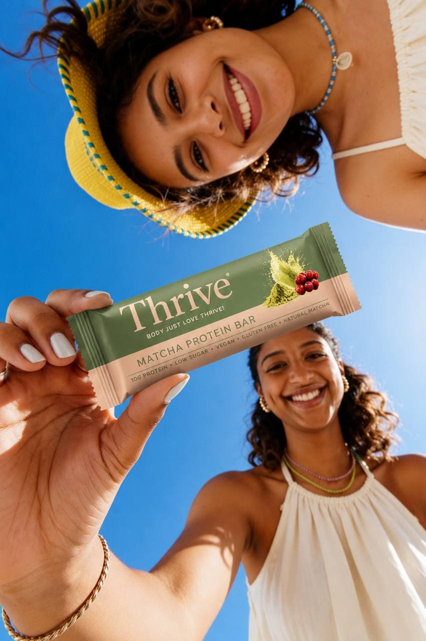 Two smiling people outdoors hold a Thrive Matcha Protein Bar, with a bright blue sky background.