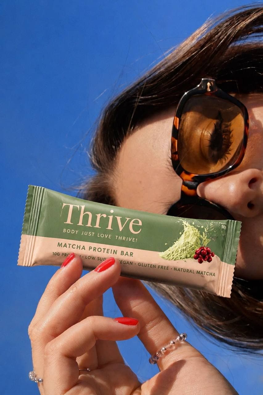 Close-up of a person holding a Thrive Matcha Protein Bar against a clear blue sky, wearing tortoiseshell sunglasses and red nail polish.