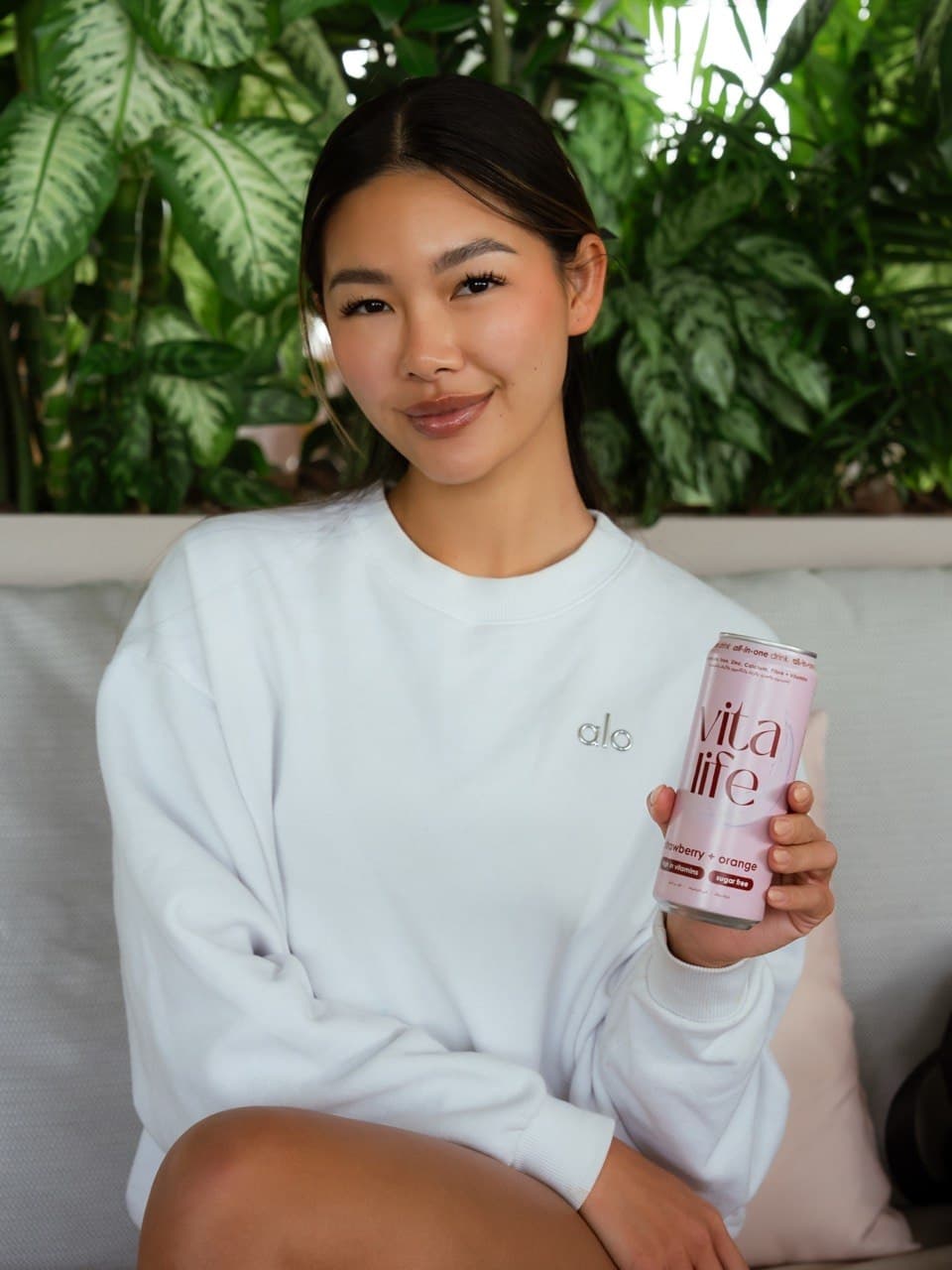 A woman in a white sweatshirt holds a can of Vita Life juice, sitting against a backdrop of green plants.